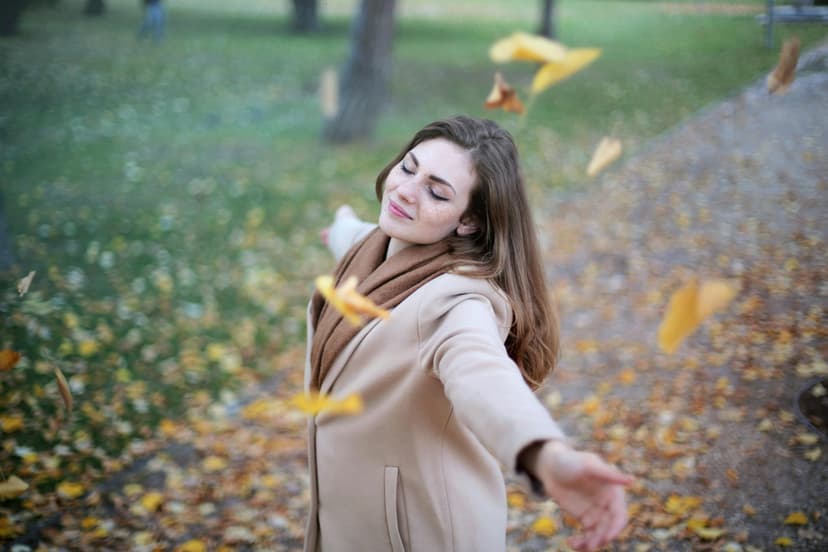 Woman enjoying a fall day with leaves falling around her in a park, symbolizing joy and freedom in nature.